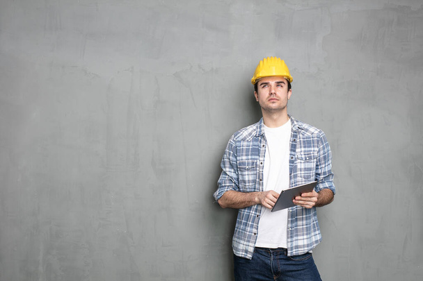 think before you act, young worker with safety helmet and a modern tablet looking for a solution to end the project that he has, standing on a concrete wall - Фото, изображение