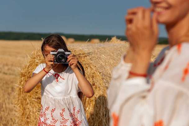красивая девочка-подросток с камерой в руках фотографирует свою улыбающуюся старшую сестру на фоне скошенного пшеничного поля и стога сена
 - Фото, изображение