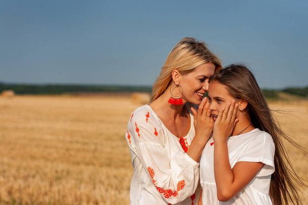 twee mooie zussen een blonde en een brunette in geborduurde shirts chatten vrolijk tegen de hemel en een tarweveld - Foto, afbeelding