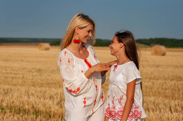 twee mooie zussen een blonde en een brunette in geborduurde shirts lachen vrolijk tegen de lucht en een tarweveld - Foto, afbeelding