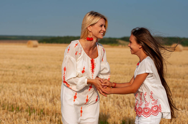zwei schöne Schwestern blond und brünett in bestickten Hemden lachen fröhlich Händchen haltend auf einem Weizenfeld - Foto, Bild