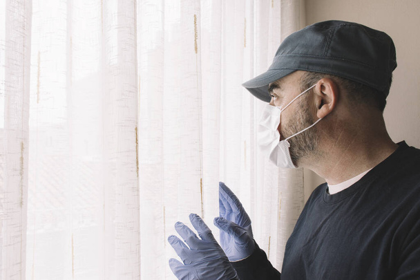 Man met gezichtsmasker die uit het raam van zijn huis kijkt terwijl hij in quarantaine zit voor ziekte. Begrippen griep, coronarivus of pandemie - Foto, afbeelding