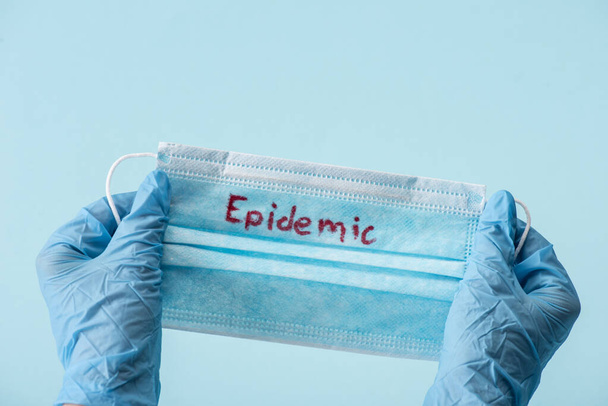 oříznutý pohled na lékaře v latexových rukavicích držícího lékařskou masku s epidemickým nápisem izolovaným na modré  - Fotografie, Obrázek