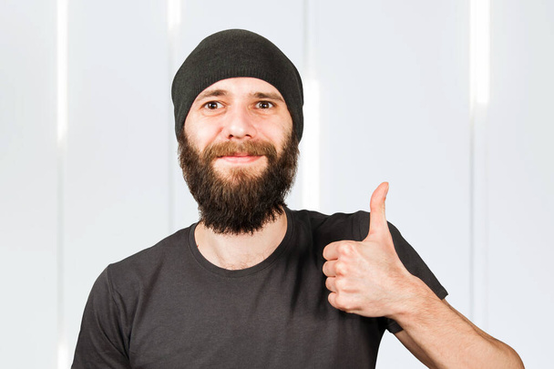 Young bearded guy in hat, smiles and show thumb up. - Zdjęcie, obraz