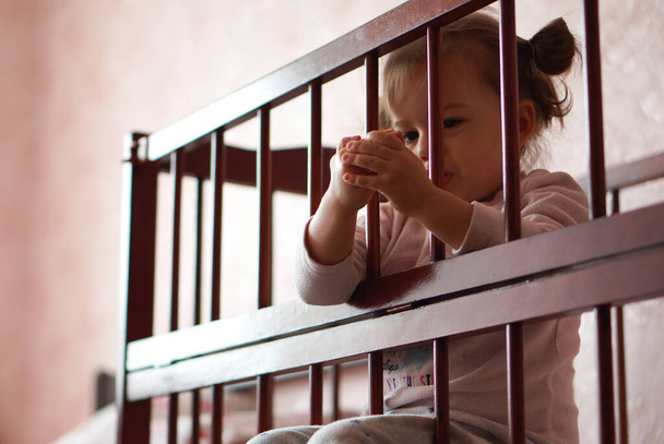 Little beautiful girl dressed in pajamas sits in a cradle. A baby in a pink pajamas sits in a cot - Photo, Image