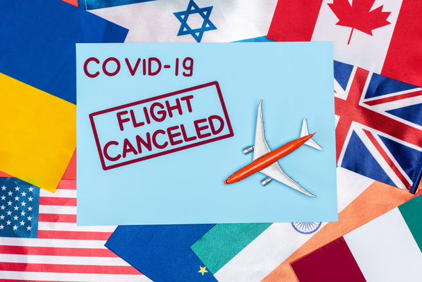 Draufsicht auf blauen Umschlag mit Covid-19 und Flugausfall-Schriftzug in der Nähe von Spielzeugflugzeug auf verschiedenen Flaggen - Foto, Bild