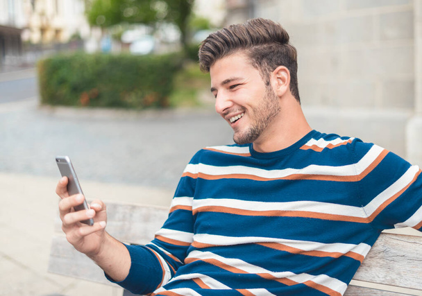 Πορτρέτο του νεαρού άνδρα χαμογελώντας στην κάμερα, ενώ τα μηνύματα σε απευθείας σύνδεση με σύγχρονο smartphone - Φωτογραφία, εικόνα