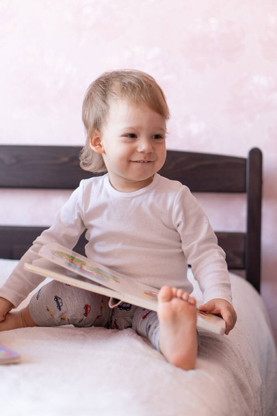 Милый блондин-мальчик читает книгу дома, сидя в постели. Маленький мальчик улыбается сидя на кровати
. - Фото, изображение