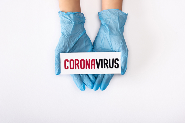 top view of doctor in latex gloves holding card with coronavirus lettering on white  - Fotó, kép