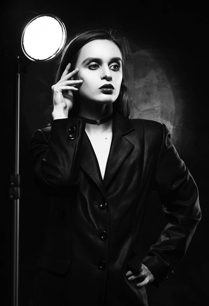 Beautiful slim girl model with red lips, wearing a black blazer, touches her face with her hand on a dark background, posing next to a light lamp. Advertising, monochrome, black and white design - Fotó, kép
