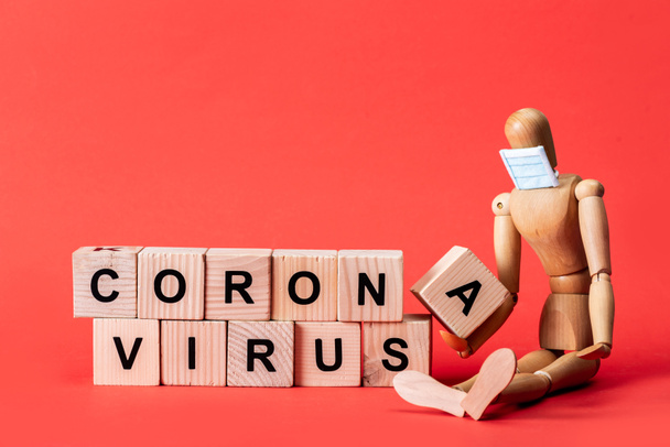 wooden doll in small medical mask near cubes with coronavirus lettering on red  - Photo, Image