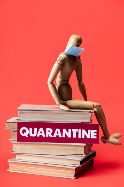 wooden doll in medical mask on books near card with quarantine lettering on red  - Foto, imagen