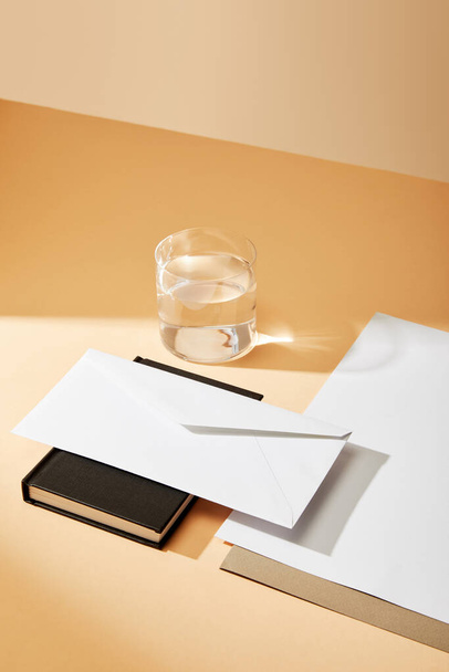 high angle view of envelope on black notebook, sheets of paper and glass of water on beige background - Photo, Image