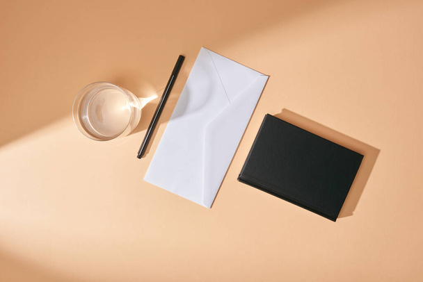 top view of envelope, pen, glass of water and black notebook on beige background - Fotó, kép