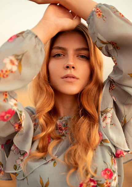 Beautiful young female with long red hair in trendy outfit with floral print looking at camera while standing against blurred autumn background - Zdjęcie, obraz
