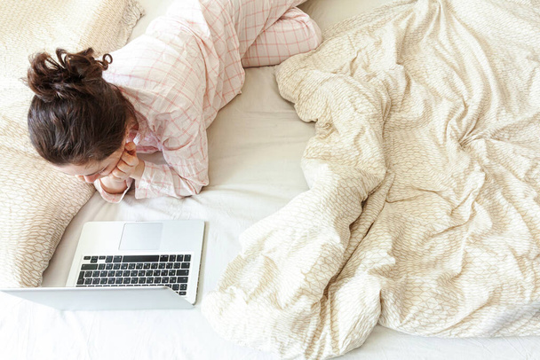 Liikkuva toimisto kotona. Nuori nainen pyjamassa istuu sängyllä kotona työskentelemässä kannettavan tietokoneen avulla. Elämäntapatyttö opiskelee sisätiloissa. Freelance liiketoiminnan karanteeni käsite
 - Valokuva, kuva