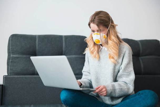 young blond woman wearing medical protection mask works in her laptop from home isolated during quarantine, virus epidemic concept. - 写真・画像