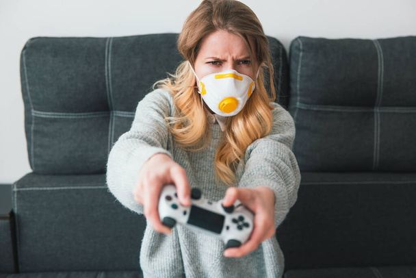 young blond woman wearing medical protection mask playing online game with joystick in her hands looks excited sitting at home during quarantine, virus epidemic concept. - Foto, imagen