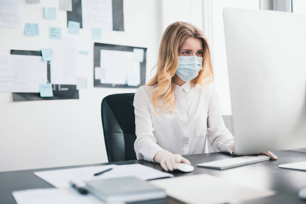 young blond woman manager in medical protection mask and gloves works in office during Covid-19 epidemy, virology concept. - 写真・画像