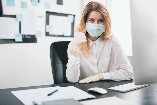 young blond woman manager in medical protection mask and gloves works in office during Covid-19 epidemy, shows like sign, virology concept. - Φωτογραφία, εικόνα