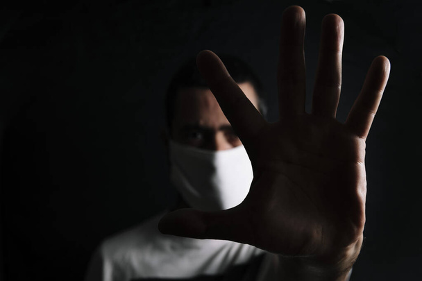 dark portrait of a man with a protective mask doing stop sign with the hand in black background, selective focus, concept of coronavirus and protection against viruses and bacteria - Photo, image