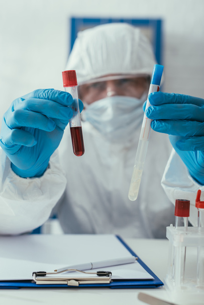 selective focus of biochemist in hazmat suit holding test tubes with blood sample and liquid - Photo, Image