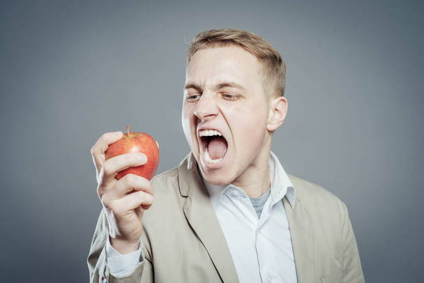 Cheerful beautiful man eating apple, isolated over gray background - Photo, Image