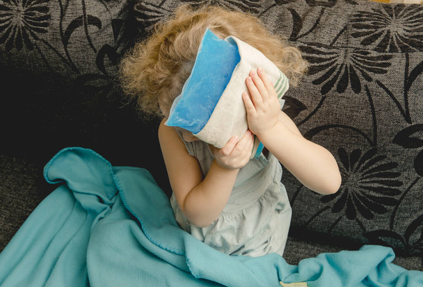 Young cute girl sit on couch in home living room and holds cold gel ice pack on head. Headache, fever or concussion concept. - Photo, Image