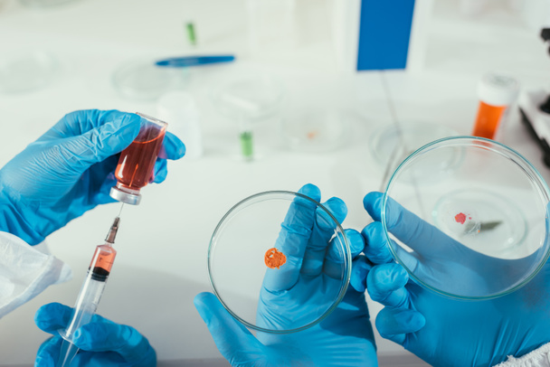 bijgesneden beeld van biochemicus die medicijnen inneemt met spuit in de buurt van collega die petrischaaltjes met biomateriaal vasthoudt - Foto, afbeelding