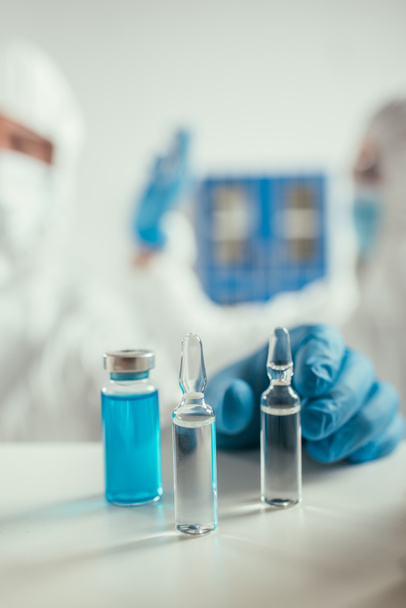 selectieve focus van biochemicus in rubberen handschoenen die ampul in de buurt van glazen container met blauwe vloeistof nemen - Foto, afbeelding