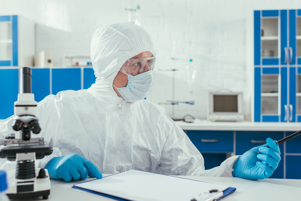 bioquímico en traje de materiales peligrosos sentado cerca del microscopio y portapapeles en el laboratorio
 - Foto, Imagen