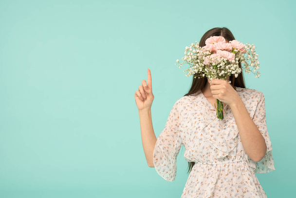 Young female in dress covered her face with spring flowers and pointing finger up, isolated over blue background - Фото, зображення
