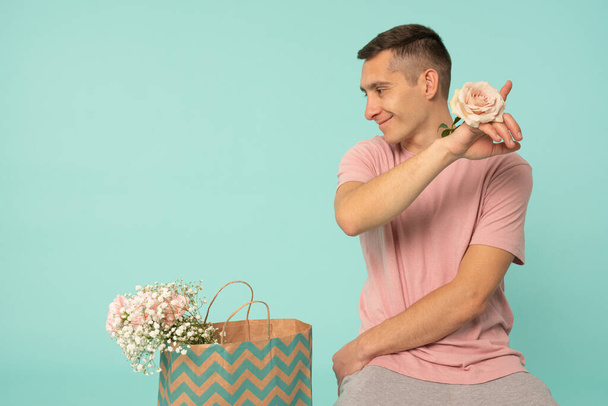 Attractive young man in pink t-shirt with rose on his hand pointing at right upper corner, sitting on the ground near the shopping bag with flowers and looking at left side - Foto, imagen