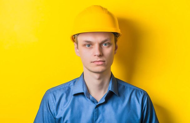 Young man close-up in yellow construction helmet and a blue shirt on a yellow background looking at the camera. Mimicry. Gesture. photo Shoot - Φωτογραφία, εικόνα