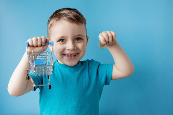 Boy in blue t-shirt is holding shopping cart and coin on blue background. Shopping and sale concept - Фото, зображення