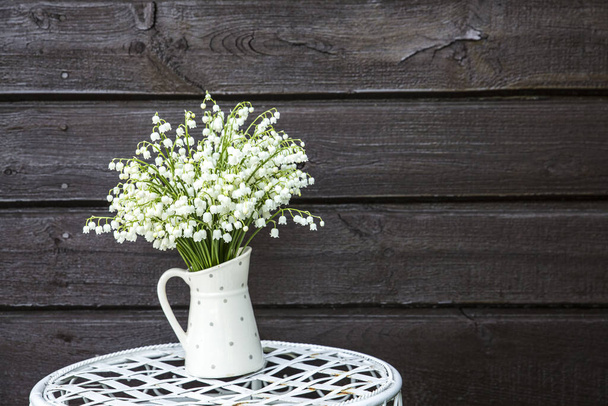Bouquet di fiori bianchi Giglio della valle (Convallaria majalis) chiamato anche: Campane di maggio, lacrime della Madonna e lacrime di Maria in un vaso bianco a forma di brocca punteggiata, all'aperto su un chiosco di fiori fantasia
. - Foto, immagini
