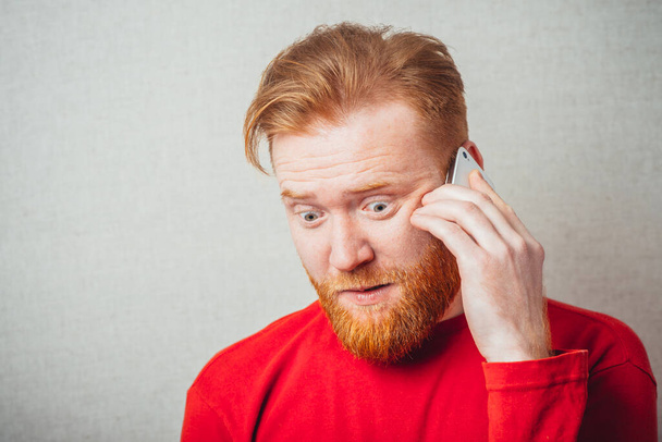 on a gray background, bearded man in a red shirt talking on the phone - Foto, Imagem
