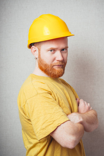 on a gray background man with a beard in a yellow shirt and yellow construction helmet folded hands - Photo, Image