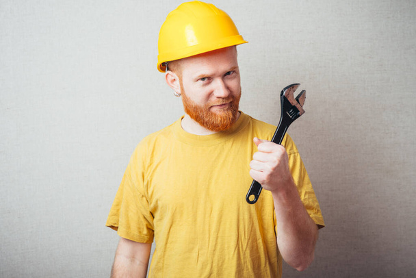 on a gray background man with a beard in a yellow shirt and yellow construction helmet holding a wrench - Foto, afbeelding