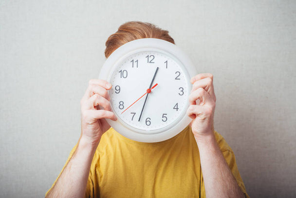 on a gray background man with a beard in a yellow shirt holding a clock and scared - Foto, afbeelding