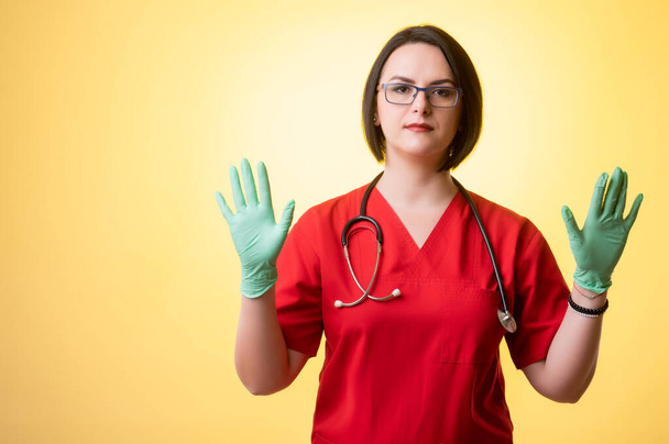 Retrato de mulher bonita médico com estetoscópio vestindo esfoliação vermelha, mostra ambas as mãos com luvas médicas posando em um fundo isolado amarelo
. - Foto, Imagem