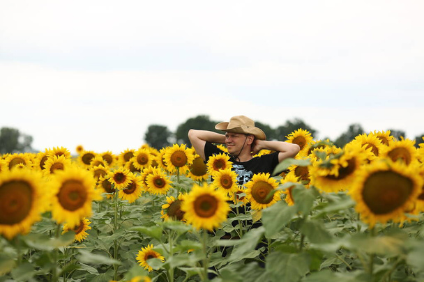 Young male farmer in a hat in a field where yellow sunflowers are blooming. Agriculture concept. Sunflower oil production. - Фото, изображение