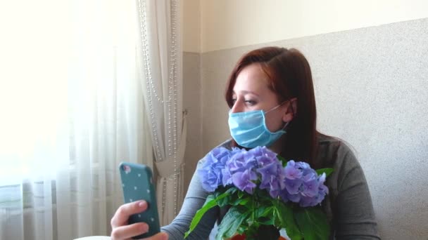 Γυναίκα με ιατρική προστατευτική μάσκα σε καραντίνα στο σπίτι. Κορίτσι που χρησιμοποιεί το κινητό τηλέφωνο, παίρνει μια selfie. Πανδημική διάθεση. Αυτοαπομόνωση κατά τη διάρκεια επιδημίας. Κορίτσι με μπλε λουλούδια ορτανσία. - Πλάνα, βίντεο