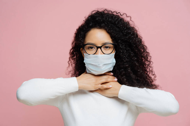 Sick curly haired woman touches neck, suffers from suffocation and shortage of breathing, wears medical mask to avoid virus infection, isolated on pink background. Coronavirus, health care concept - Photo, Image