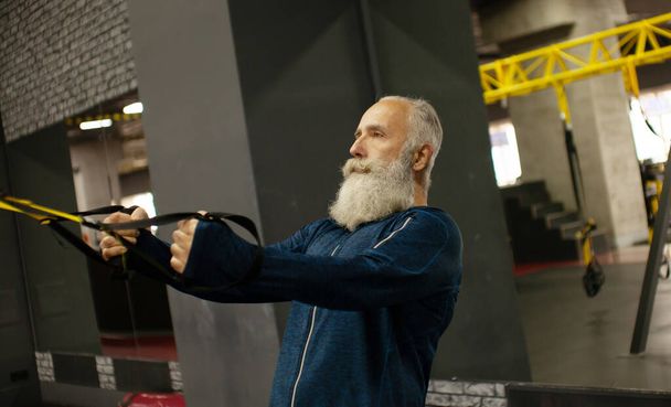 Bearded senior sportsman training with trx resistance bands in gym. - Foto, Imagen