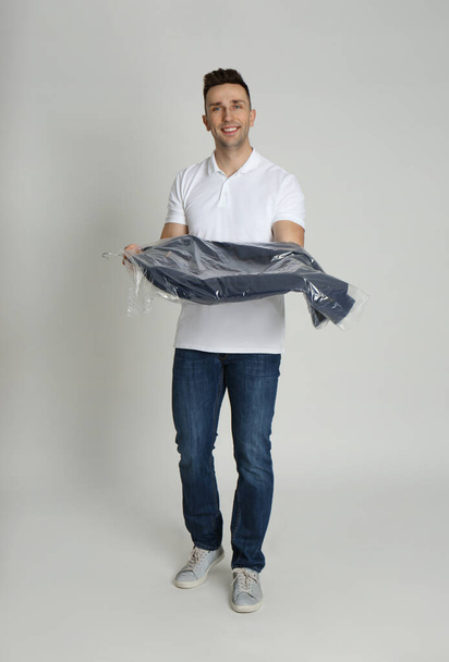 Man holding hanger with jacket in plastic bag on light grey background. Dry-cleaning service - Foto, afbeelding