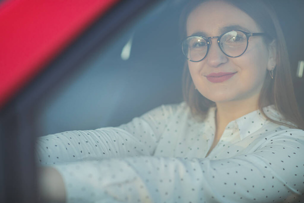 Portrait of a girl in glasses through the window of a car. Positive looking girl. Beautiful girl sitting in the new red car. - Foto, Imagen