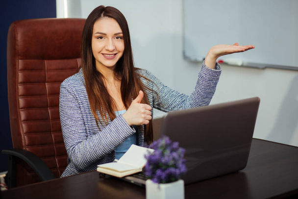 Χαρούμενος εργαζόμενος δακτυλογραφεί στο laptop και κοιτάζοντας μακριά με ένα φωτεινό χαμόγελο. Κάθεται σε ένα γραφείο σε ένα γραφείο. Πορτρέτο. - Φωτογραφία, εικόνα