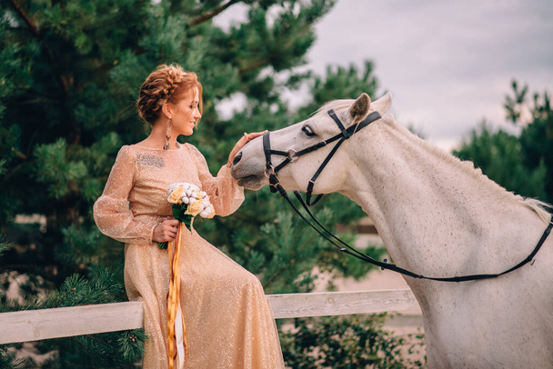 Молодая невеста в золотом платье гладит голову белой лошади
 - Фото, изображение
