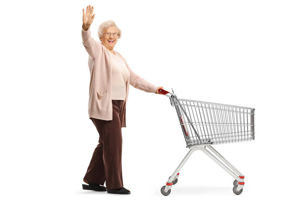 Full length shot of a senior lady waving and pushing an empty shopping cart isolated on white background - Fotoğraf, Görsel